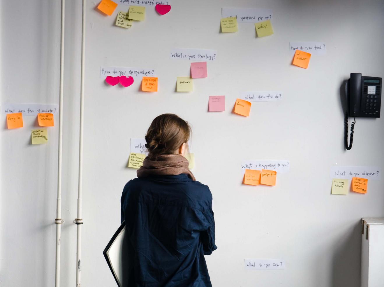 Eine Frau betrachtet eine Wand, an der Post-Its mit Fragen kleben.