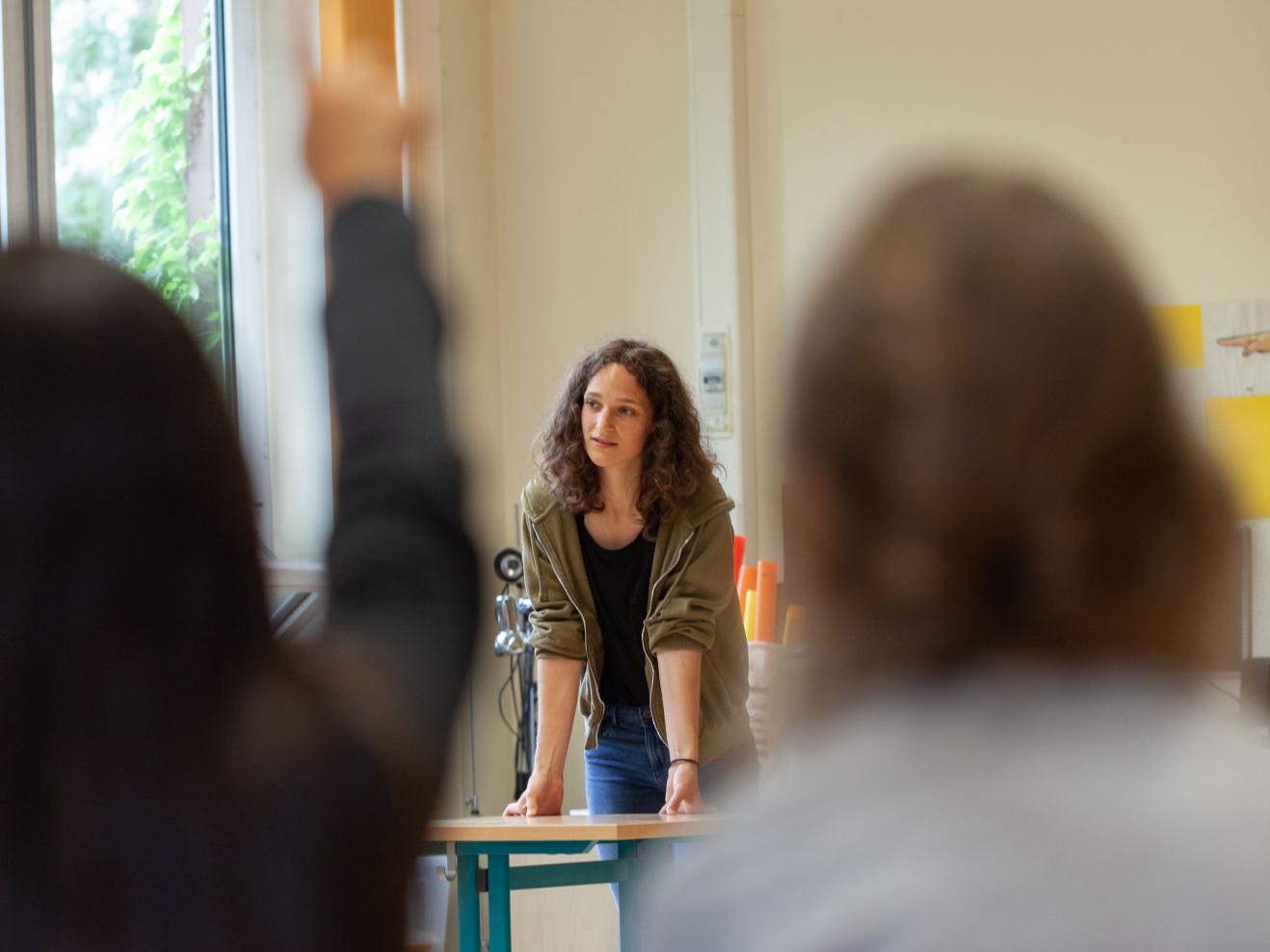 Eine Studentin stützt sich auf einem Tisch ab. Unscharf im Vordergrund meldet sich ein Kind