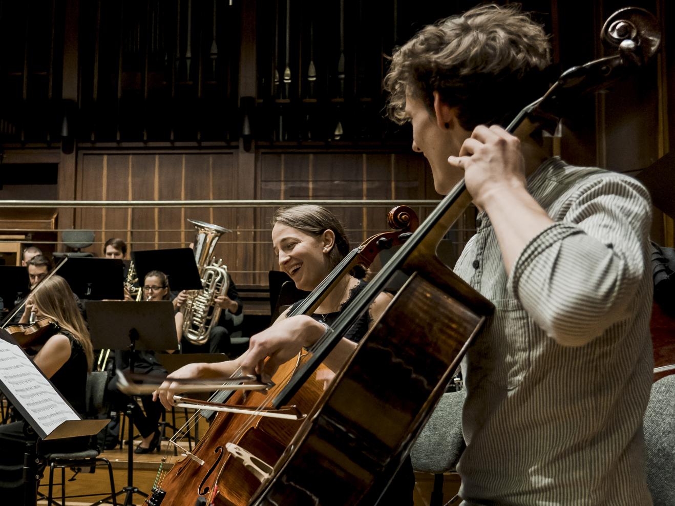 Eine Cellistin und ein Cellist in der Orchesterprobe, lachend einander zugewandt.