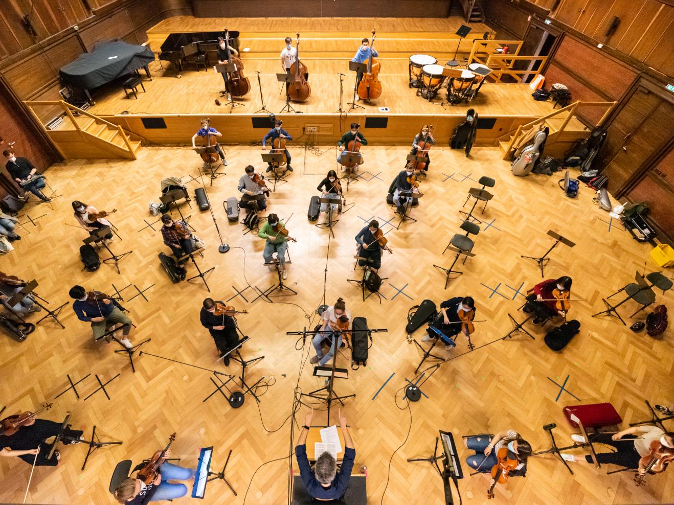 Blick aus der Vogelperspektive auf das Hochschulorchester, das mit Abstand im gesamten Großen Saal verteilt sitzt. Auf dem Boden kleben schwarze Markierungen.