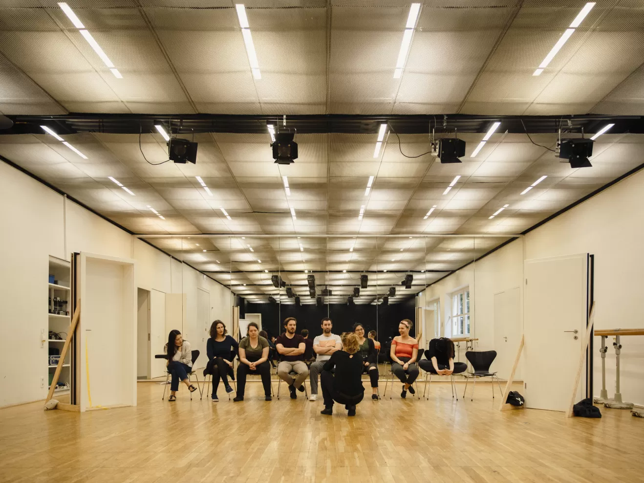 Acht Gesangsstudierende sitzen nebeneinander in einer Stuhlreihe im Opernstudio vor ihnen hockt eine Person