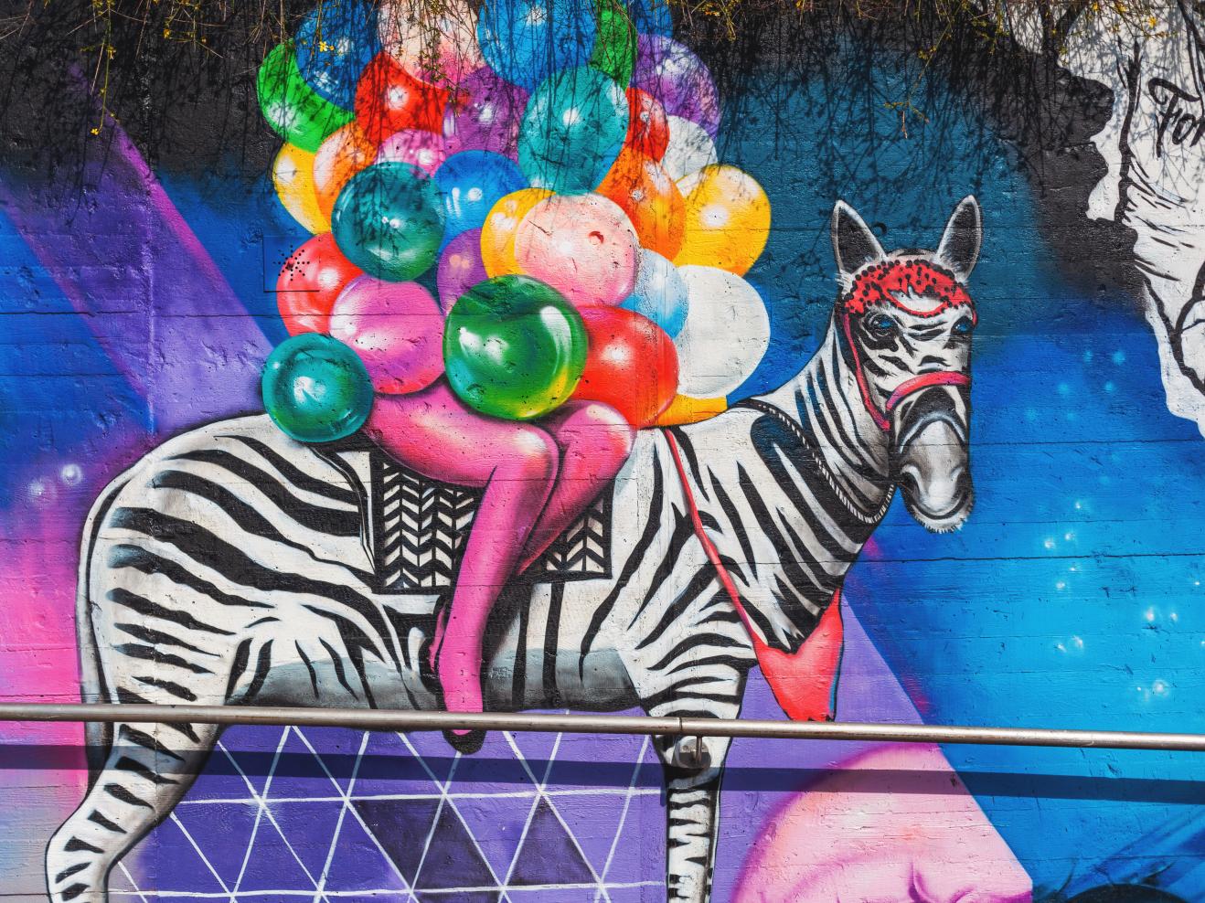 Buntes Graffiti im Stadtbild: Eine Person auf einem Zebra, die Arme übervoll mit bunten Luftballons.