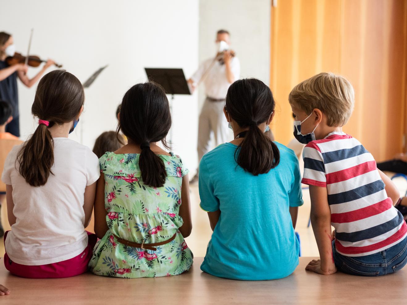 Vier Kinder sitzen mit dem Rücken zum Betrachter und hören zwei Musikern zu.