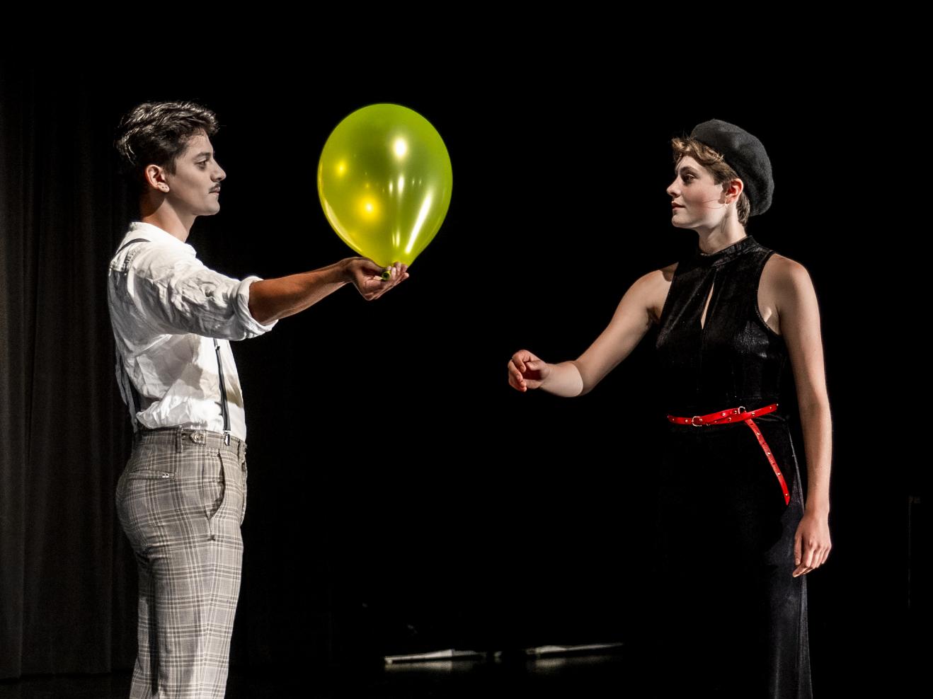 Zwei Personen auf der Bühne, eine reicht der anderen einen aufgeblasenen gelben Luftballon.