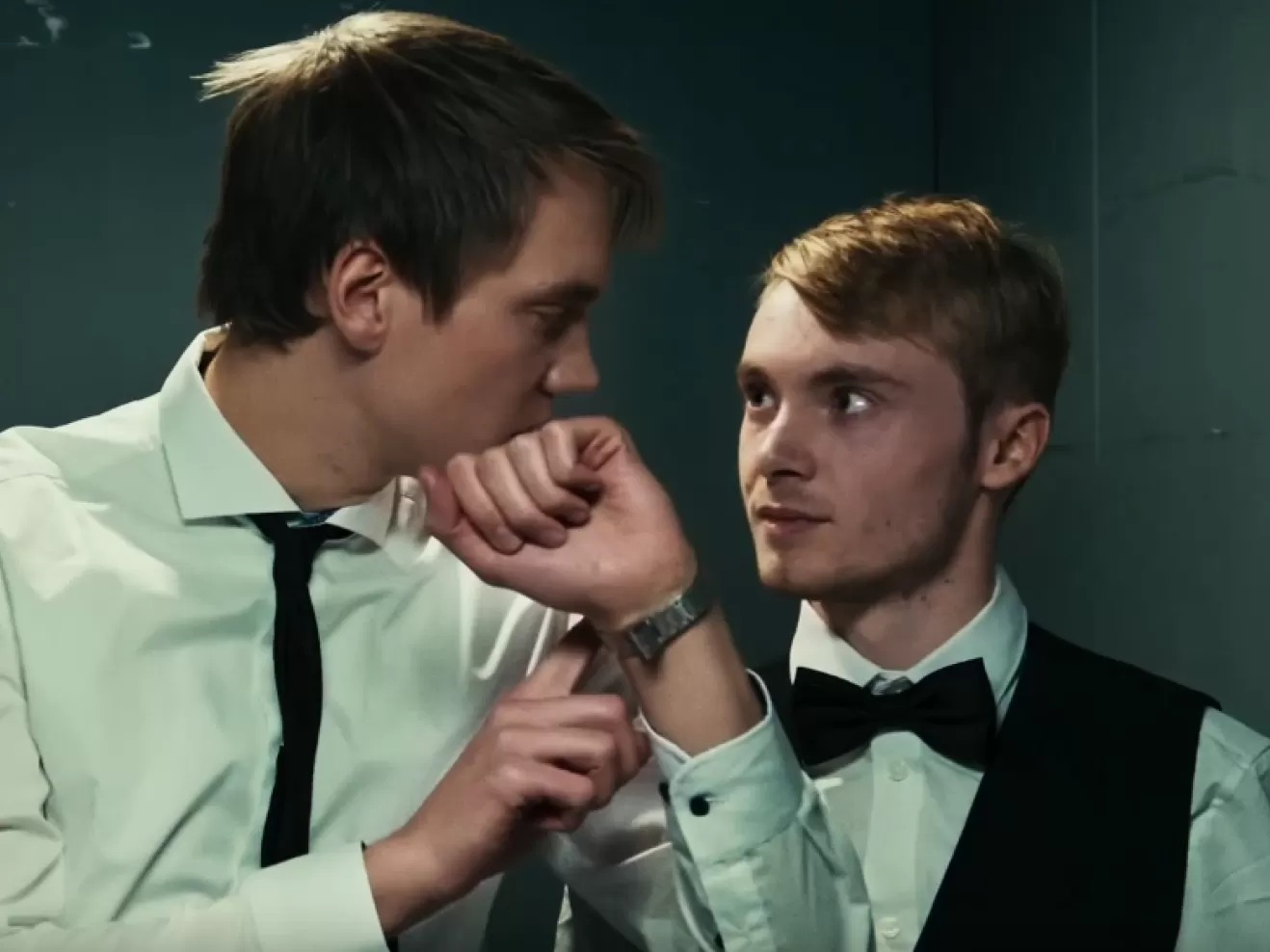 Zwei Männer im Fahrstuhl. Der eine zeigt auf seine Armbanduhr.