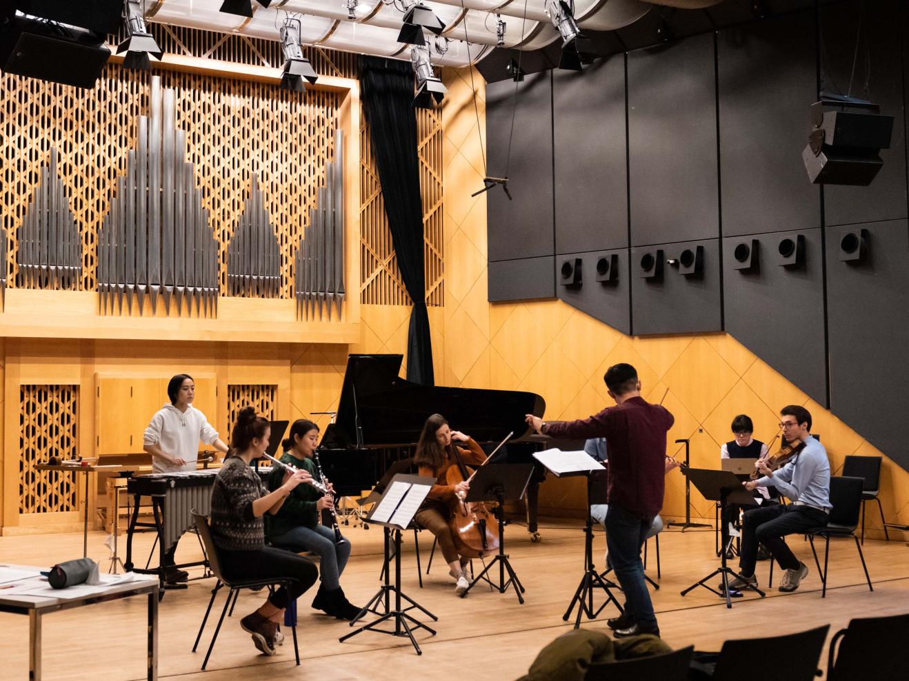 Ensemble mit 7 Musiker*innen auf der Bühne, im Vordergrund sieht man den Dirigenten von hinten.