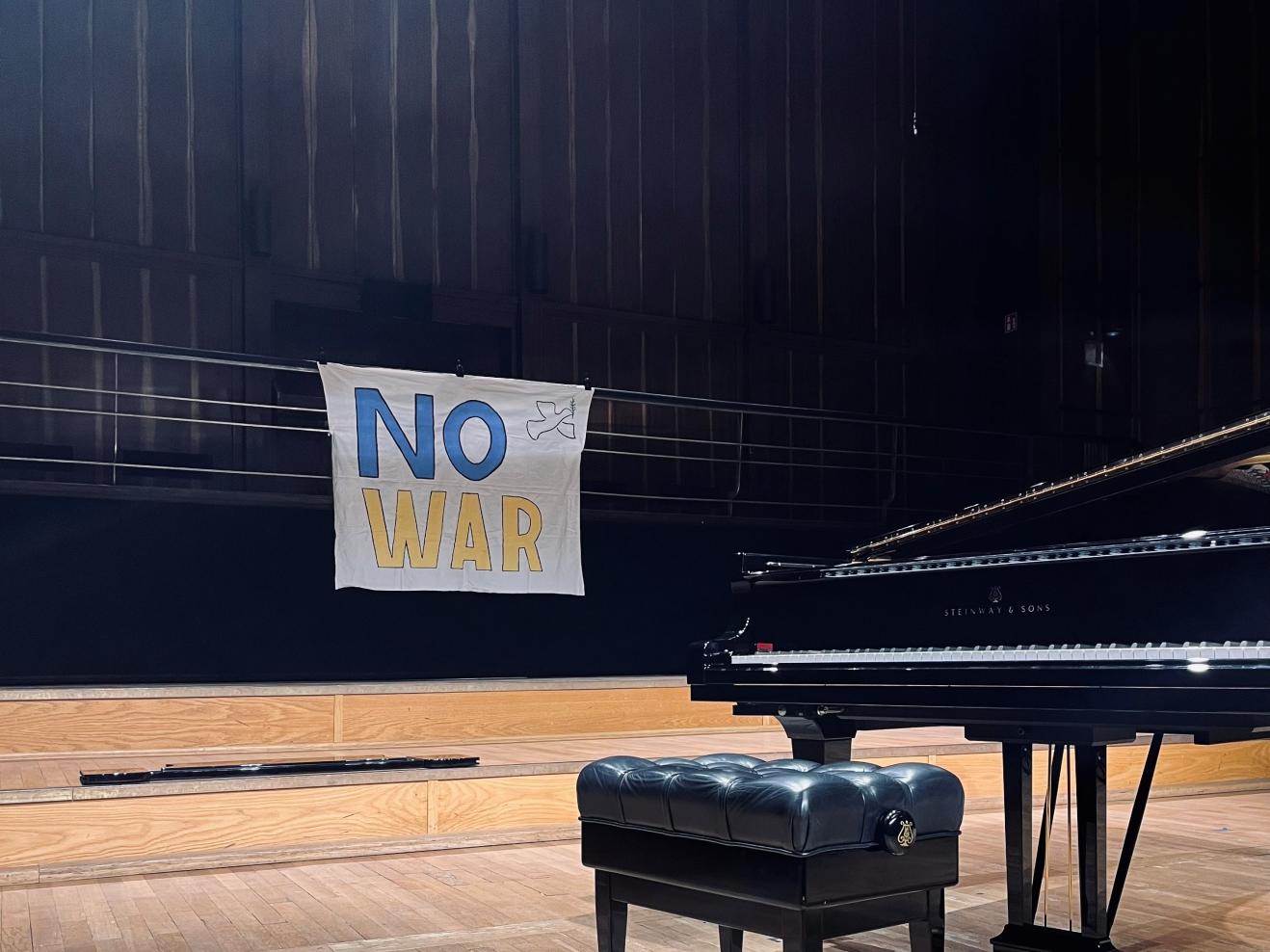 Ein Transparent mit der Aufschrift "No War" in gelb und blau hängt hinter der Bühne im Großen Saal der HfMDK, im Vordergrund ein Konzertflügel