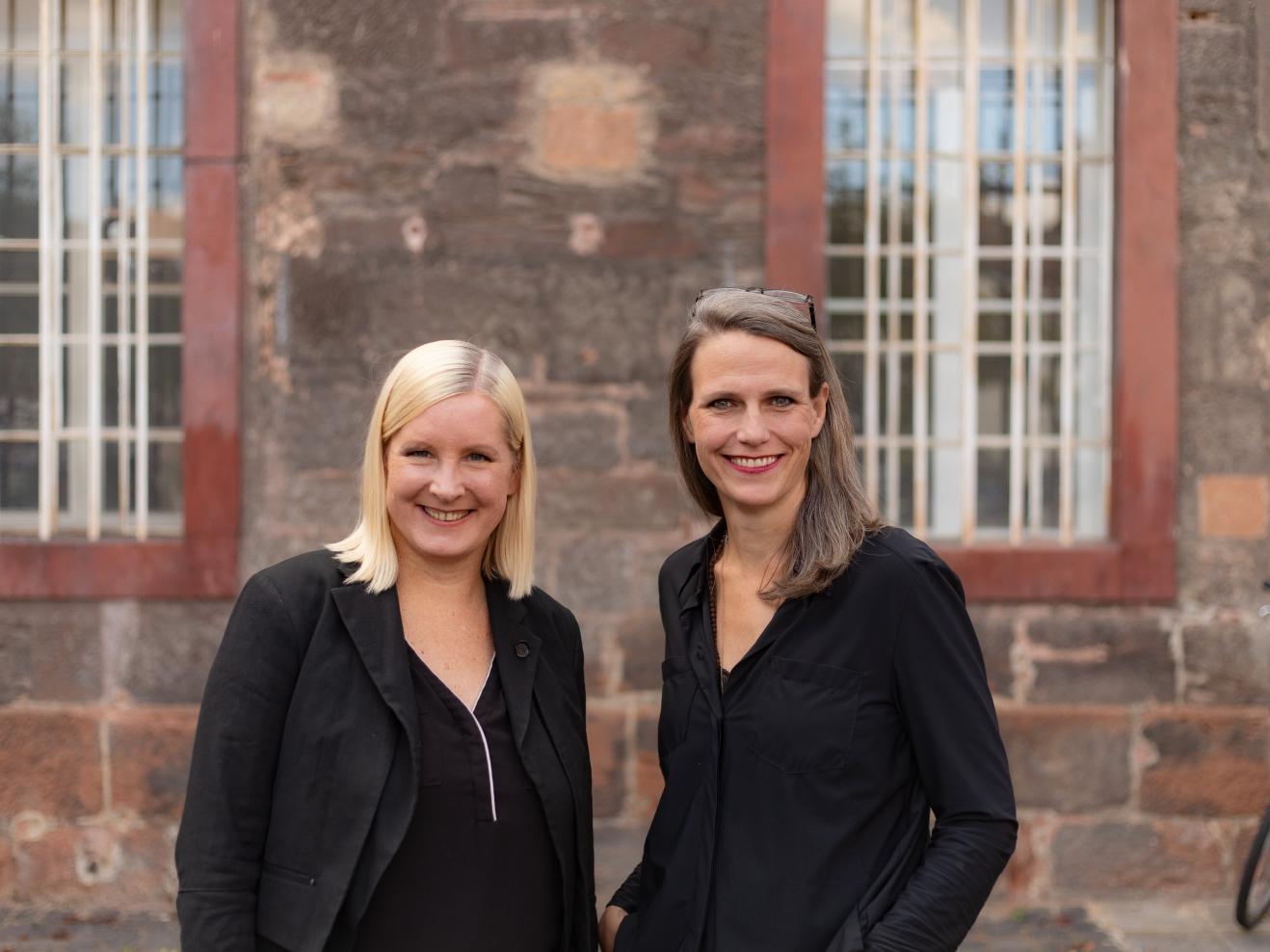 Eva Lange und Carola Unser stehen vor der rötlichen Steinwand eines Gebäudes.