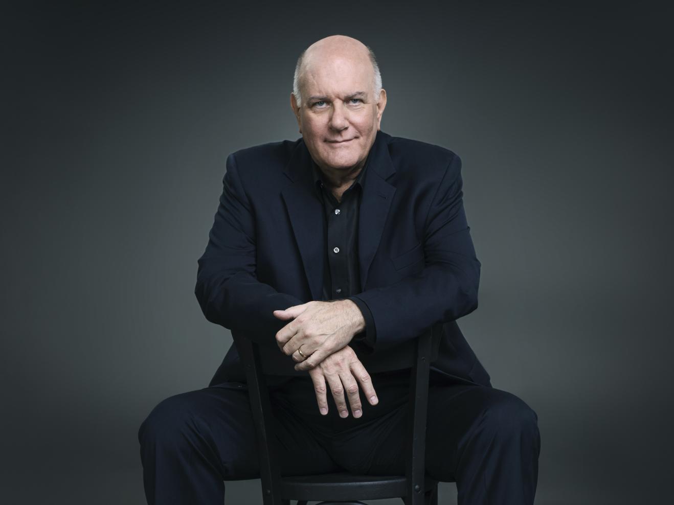Porträt von Jim McNeely, der vor einer dunkelgrauen Rückwand auf einem Stuhl sitzt.
