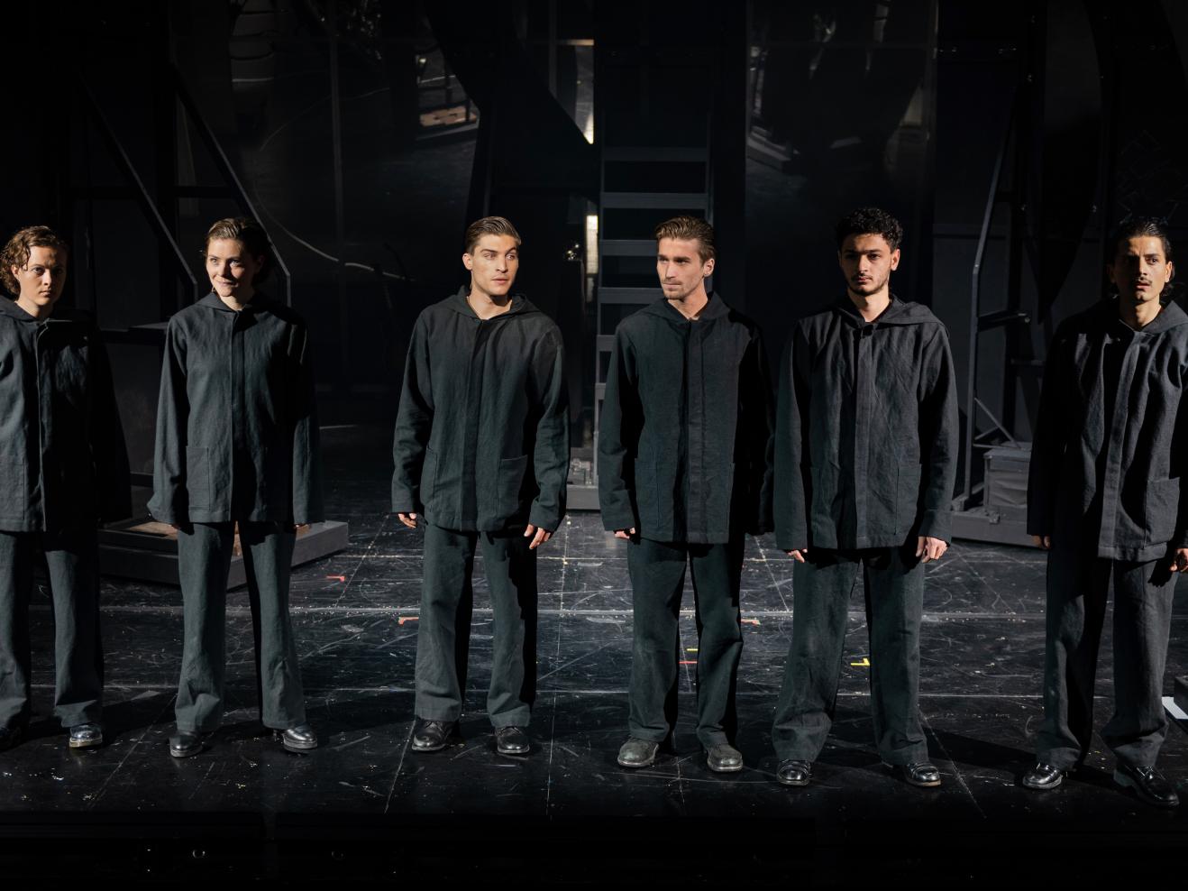 Sechs Schauspielende stehen in schwarz gekleidet nebeneinander auf einer dunklen Bühne.