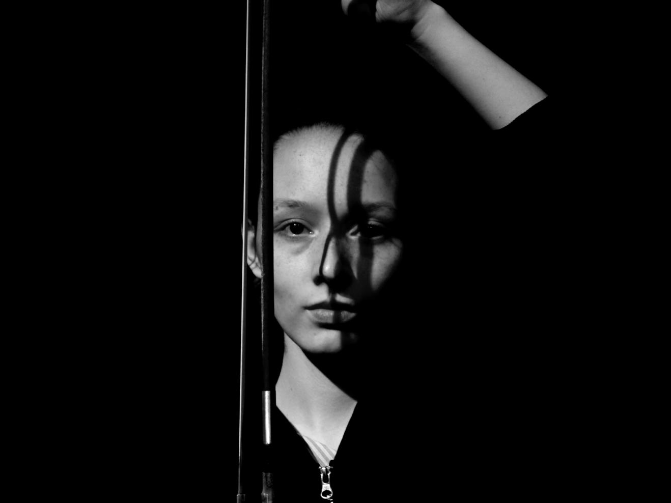 Schwarz-Weiß-Nahaufnahme einer jungen Frau, die einen Cello-Bogen hält, der einen Schatten auf ihr Gesicht wirft