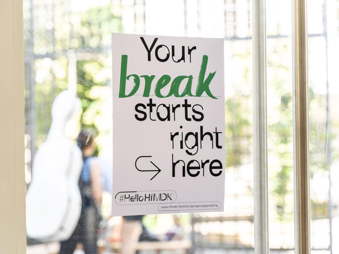 Plakat mit der Aufschrift "Your break starts right here" an der Glastür zum Innenhof der HfMDK