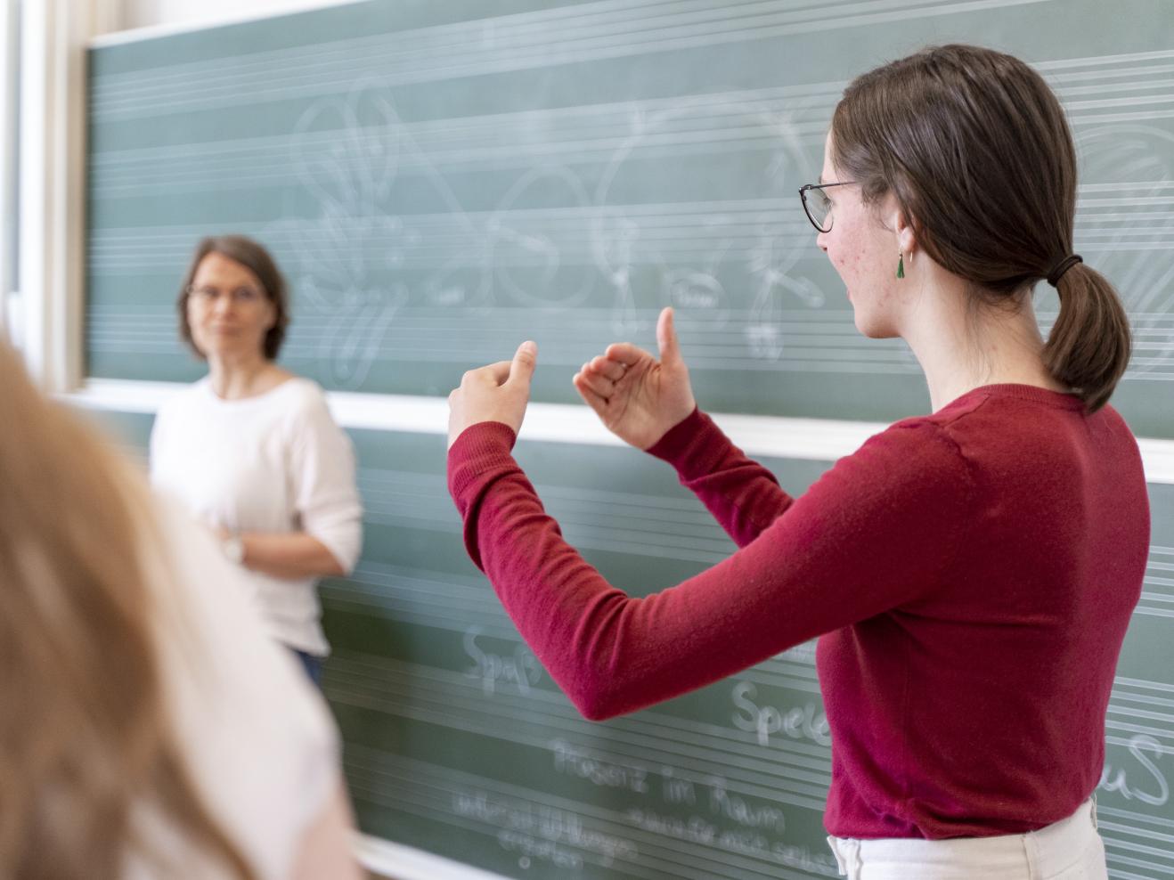 Eine Studentin im roten Pulli steht vor einer Tafel mit Notenlinien und erläutert etwas, dabei gestikuliert sie mit den Händen.