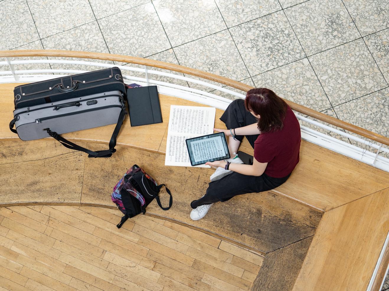 Eine Studentin, von oben gesehen, sitzt im Foyer auf der Bank mit einem Tablet, auf dem Noten zu sehen sind und einer aufgeklappten Partitur.