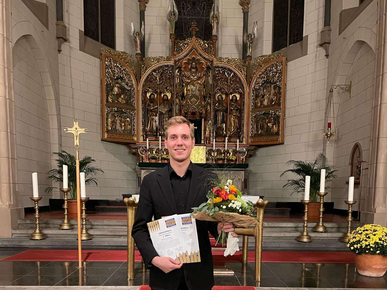 Manuel Pschorn im Anzug in einer Kirche mit Blumenstrauß und zwei Urkunden in den Händen