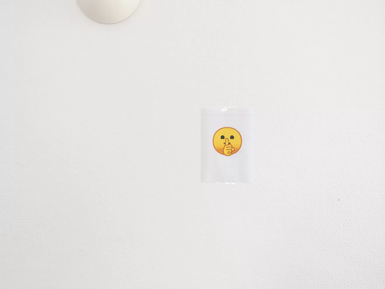 Ein Zettel mit einem gelben Emoji mit Finger auf dem Mund hängt an einer weißen Wand