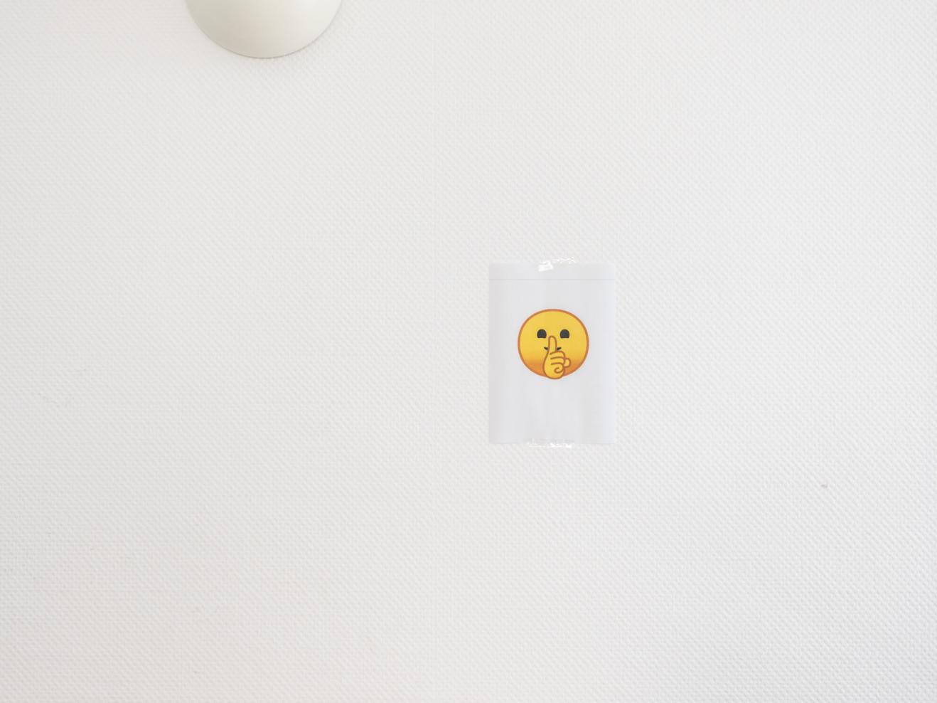 Ein Zettel mit einem gelben Emoji mit Finger auf dem Mund hängt an einer weißen Wand