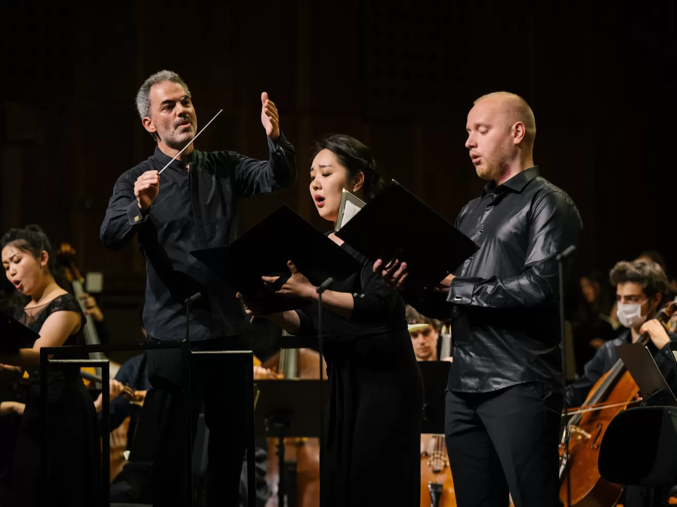Eine Sängerin und ein Sänger im Orchesterkonzert, im Hintergrund der Dirigent, der sie leitet.