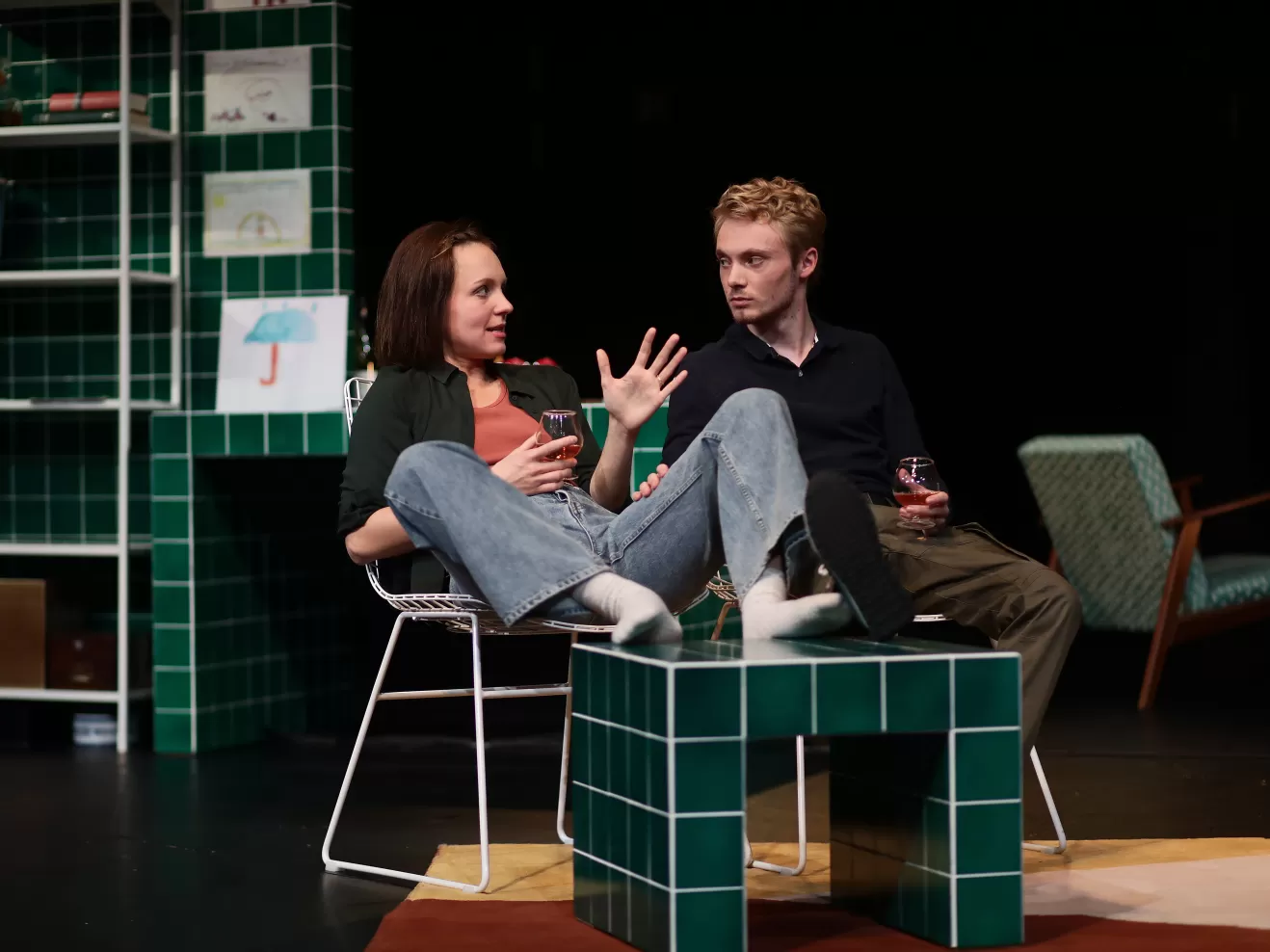 Ein Schauspieler und eine Schauspielerin sitzen Stühlen mit je einem Glas Schnaps in der Hand. Sie unterhalten sich.
