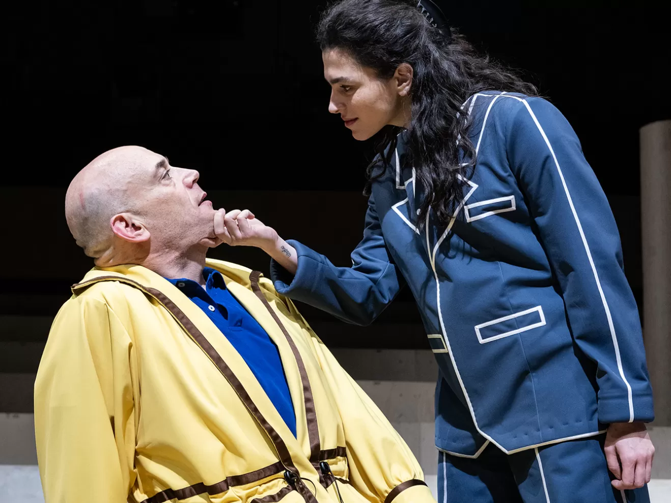 Schauspielerin im blauen Anzug hält das Kinn eines anderen Schauspielers fest.