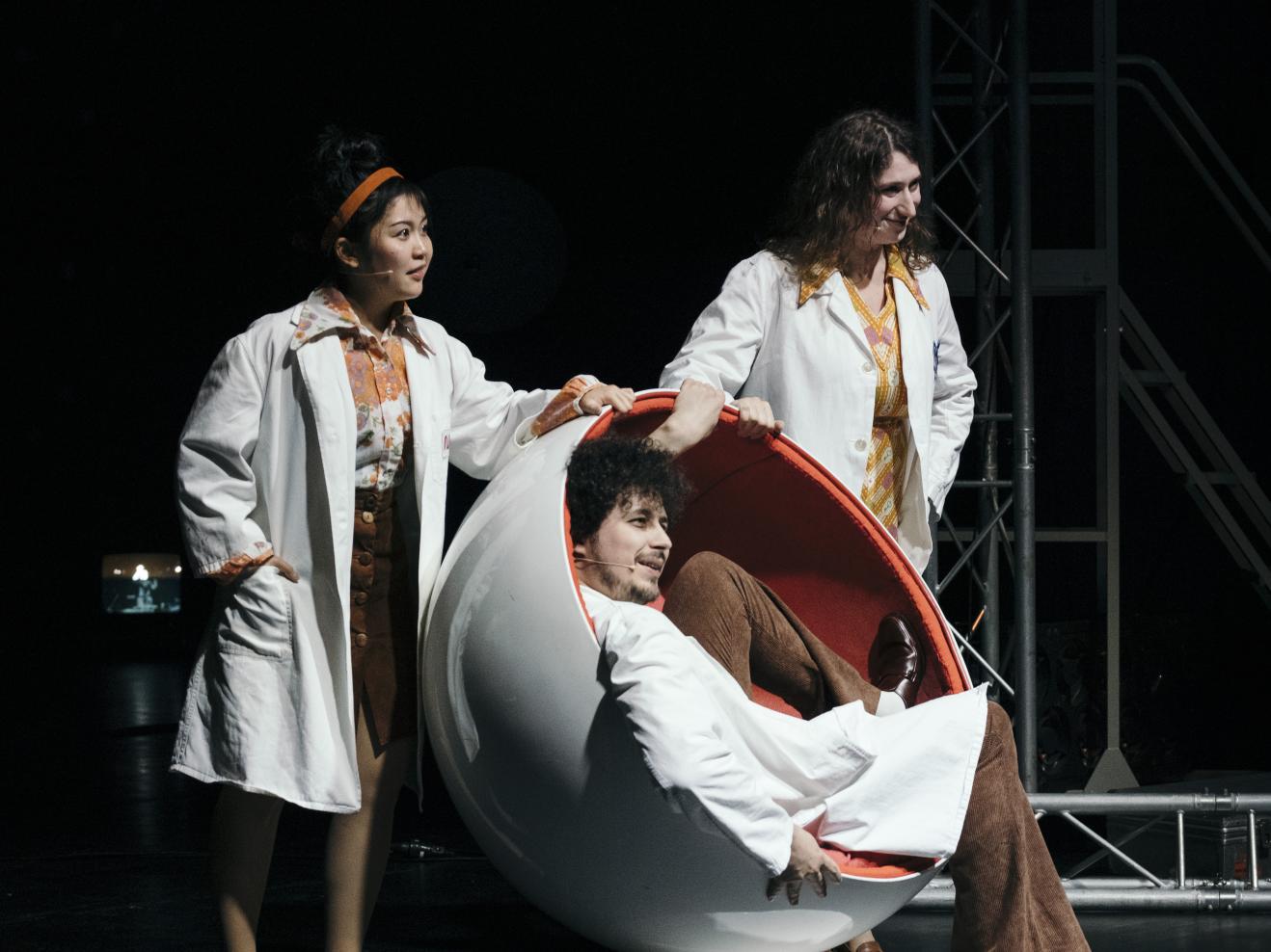 Drei Darsteller*innen in weißen Kitteln auf einer dunklen Bühne, die Person vorne sitzt in einem runden Sessel.