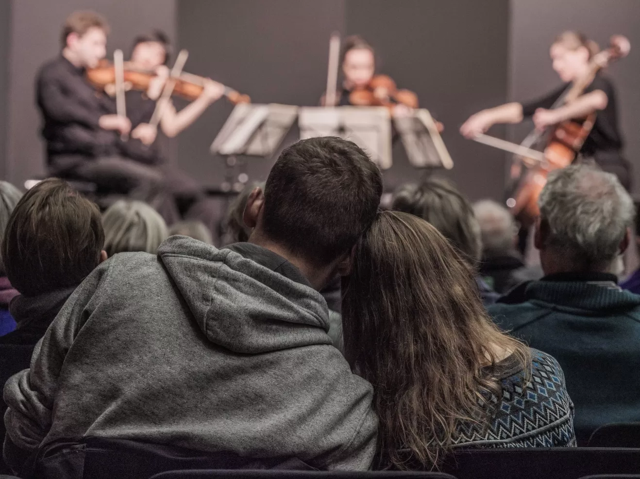 Zwei sitzende Personen lehnen ihre Köpfe aneinander. Im Hintergrund eine Bühne mit einem Streichquartett.