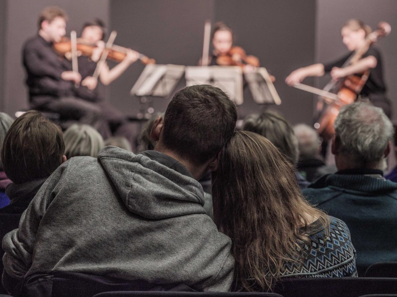 Zwei sitzende Personen lehnen ihre Köpfe aneinander. Im Hintergrund eine Bühne mit einem Streichquartett.