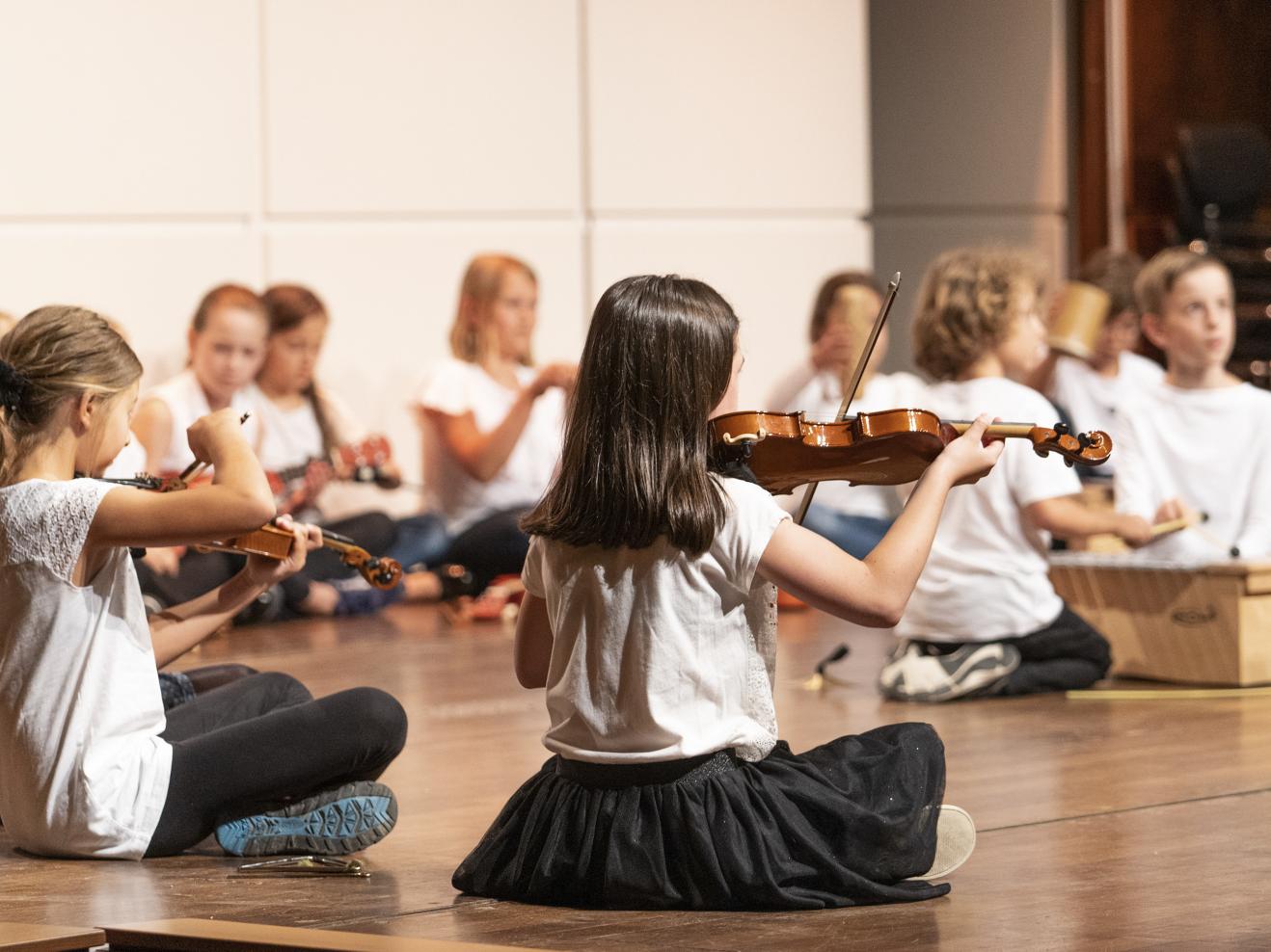Grundschüler*innen sitzen mit ihren Instrumenten auf der Bühne in der Alten Oper. Im Vordergrund ein Mädchen mit einer Geige in der Hand.