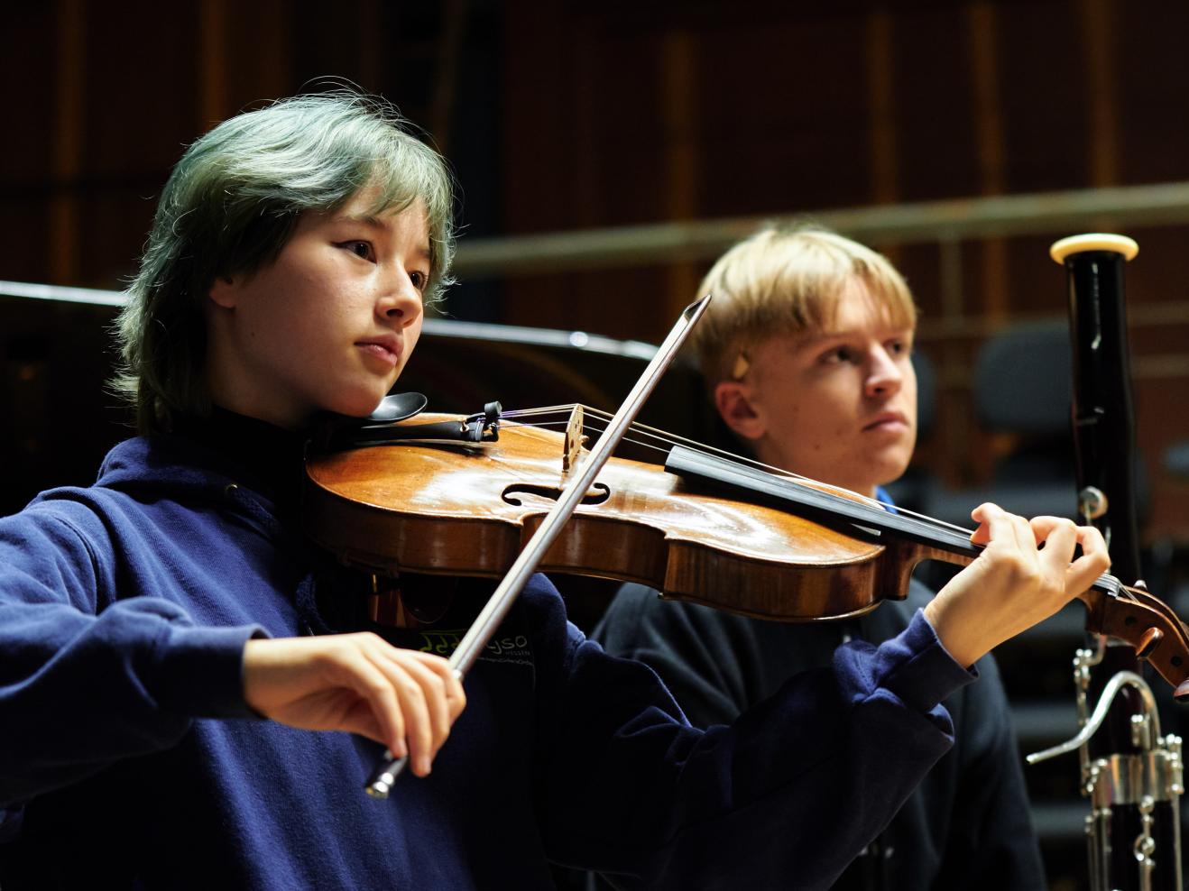 Eine Person spielt Viola, daneben sitzt im Hintergrund eine Person mit Fagott.