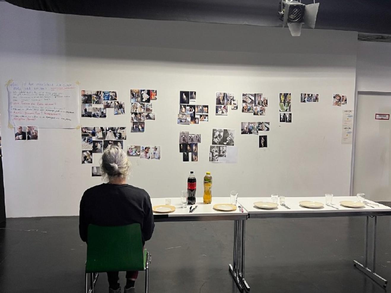 Mann von hinten sitzend vor zwei aneinandergestellten Tischen, auf diesen stehen leere Teller, Gläser und zwei Getränkeflaschen. Er blickt auf eine Wand mit vielen Fotos und einigen Notizen..