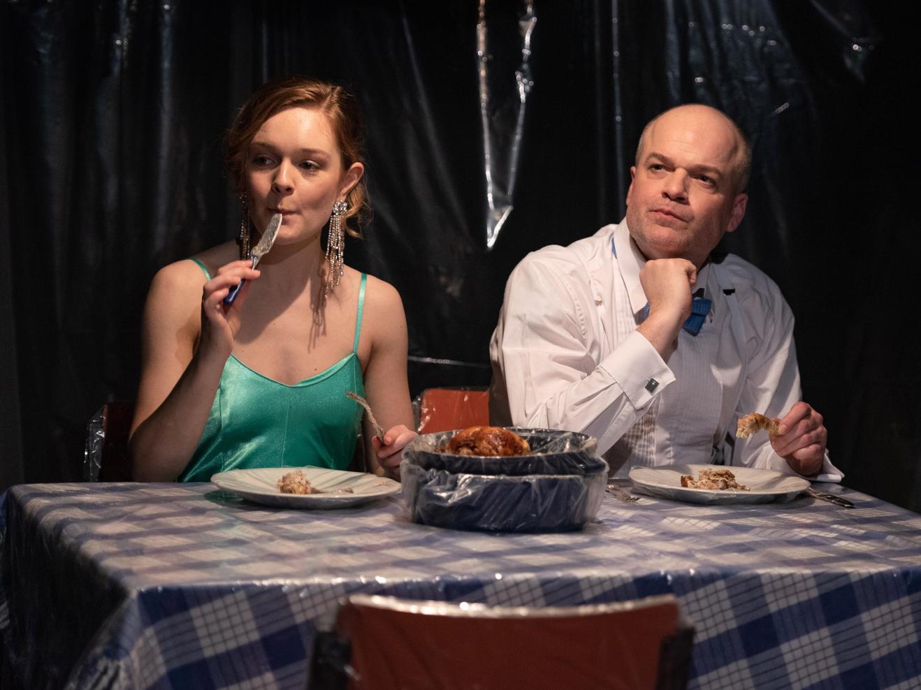Eine Frau und eine Mann, beide elegant gekleidet, sitzen an einem Tisch und essen Hähnchen.