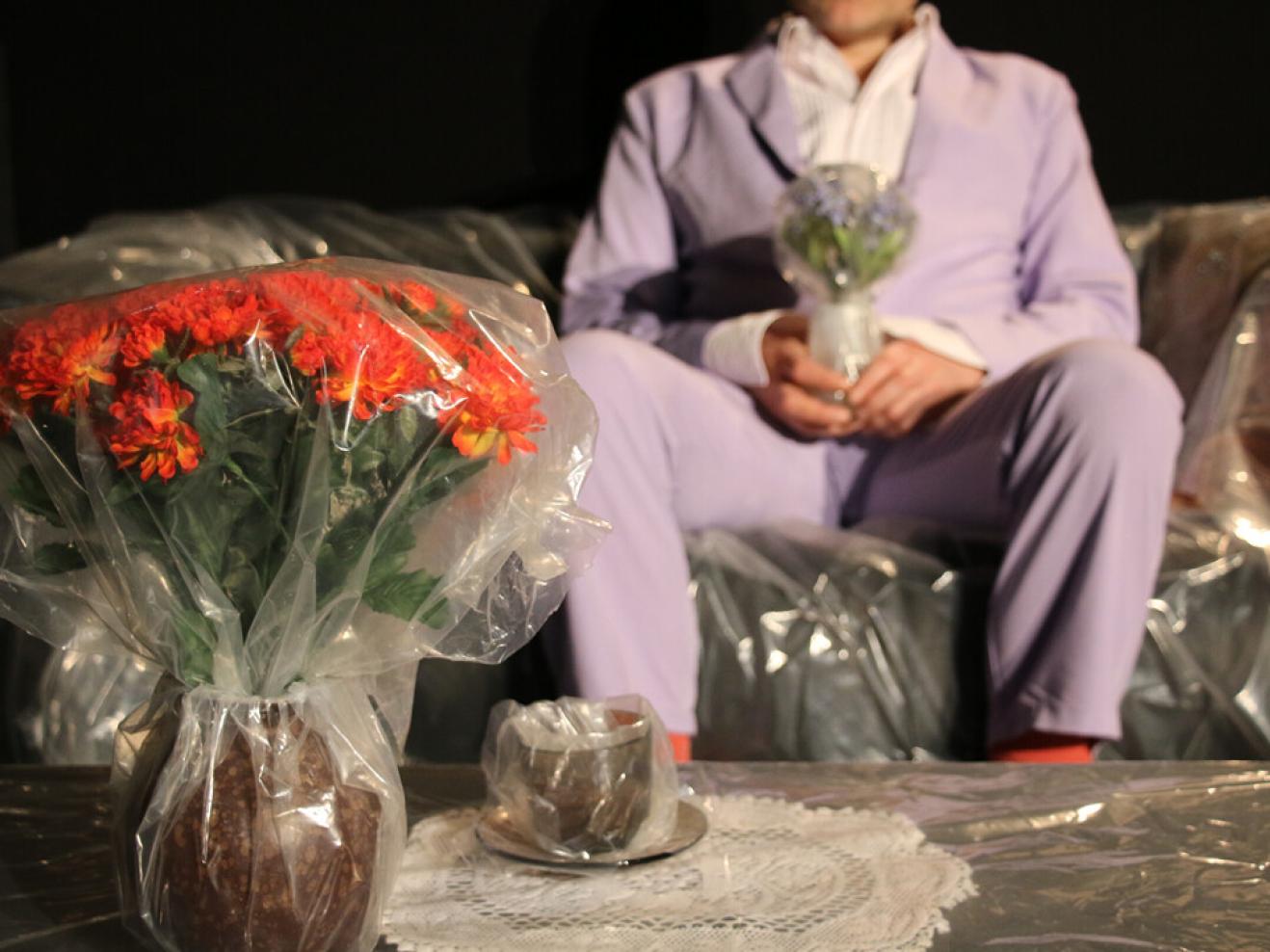In Folie verpackte Blumen stehen in einer Vase, dahinter sitzt ein Mann im Anzug.