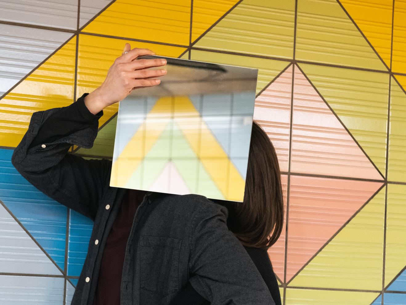 Zwei Personen in schwarz gekleidet, sie stehen eng beieinander, die Gesichter mit quadratischen Spiegeln verdeckt, die die umliegenden bunten Kacheln der U-Bahnstation Grüneburgweg reflektieren.