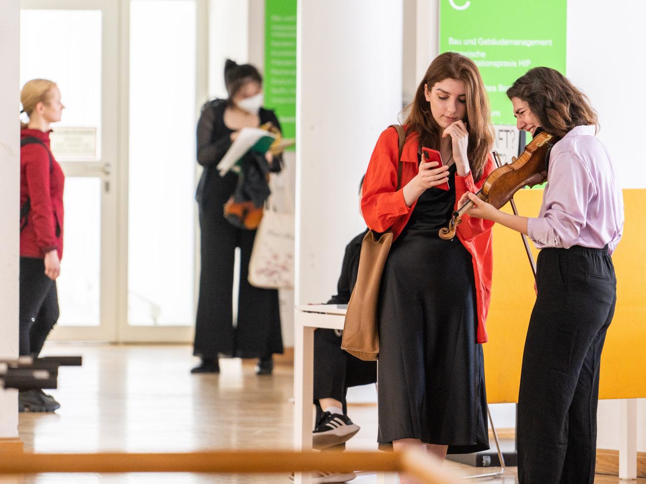 Zwei Studentinnen, von denen eine eine Geige in der Hand hält, beugen sich über ein Smartphone.