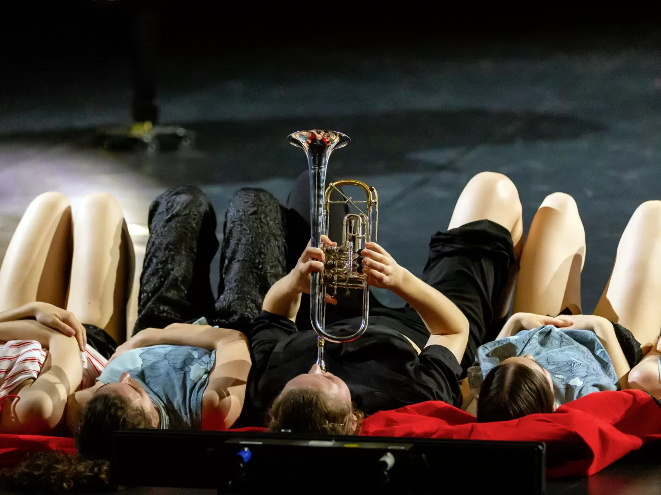 Fünf Personen liegen auf der Bühne auf dem Boden. Die Person in der Mitte spielt Trompete.