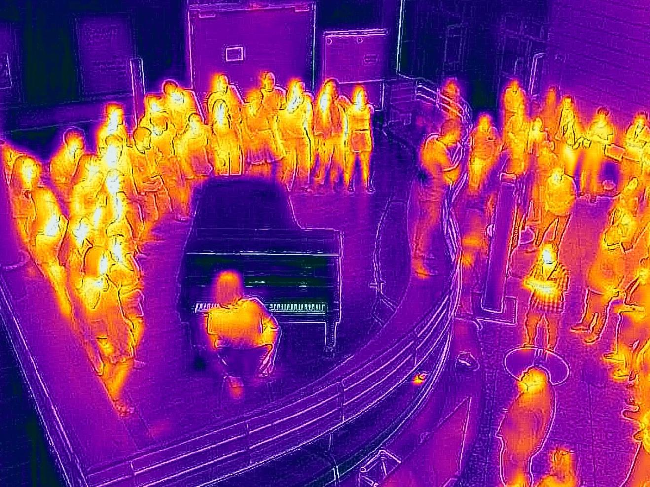 Thermografie-Aufnahme eines Konzerts im Foyer der HfMDK mit Menschen, die um einen Flügel herumstehen.