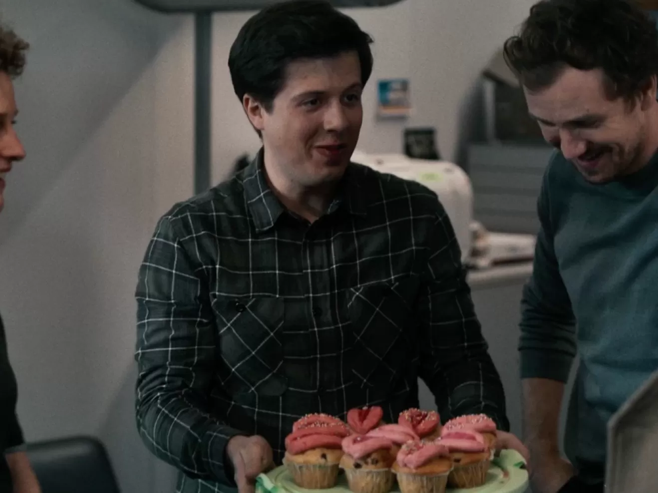 Ein Mann mit Vulva-Cupcakes auf einem Teller sowie zwei weitere Personen in eniem Büro.