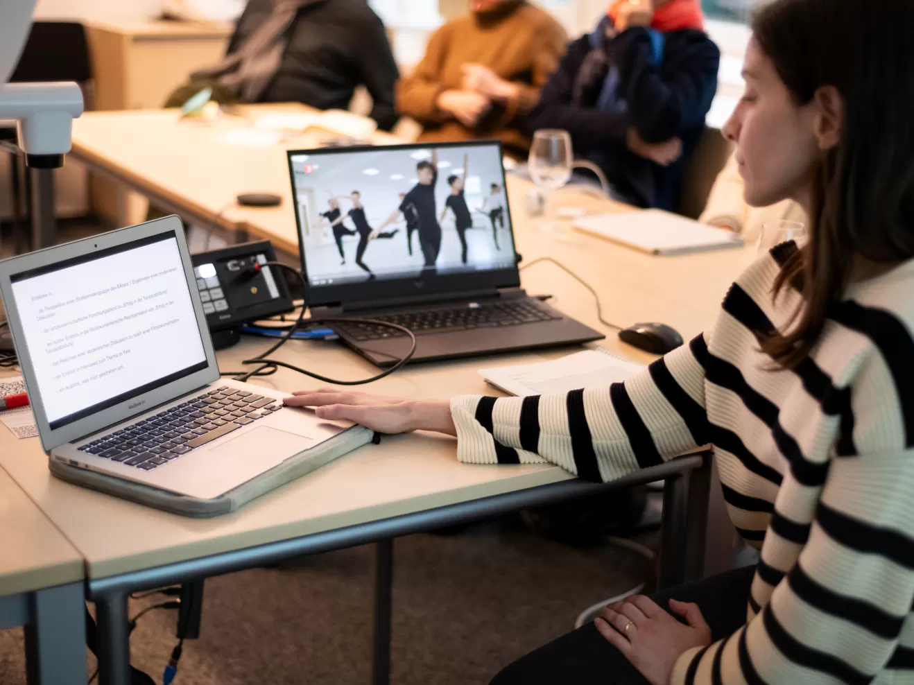Im Vordergrund rechts sehen wir eine Frau im Profil, die sitzt und auf zwei Laptops links von ihr blickt. Ein Laptop zeigt Text, der andere das Still eines Tanzvideos. Im Hintergrund sind unscharf Zuhörende zu erkennen.