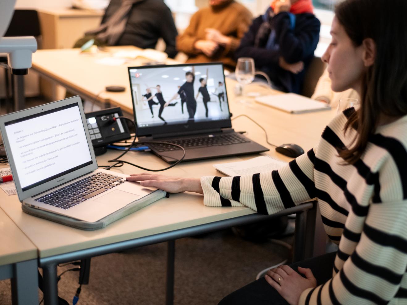 Im Vordergrund rechts sehen wir eine Frau im Profil, die sitzt und auf zwei Laptops links von ihr blickt. Ein Laptop zeigt Text, der andere das Still eines Tanzvideos. Im Hintergrund sind unscharf Zuhörende zu erkennen.
