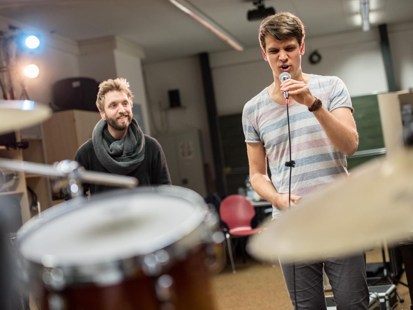 Ein Band-Coaching: Hinter einem Schlagzeug schaut ein Coach lächeln einem Studenten beim Singen zu.