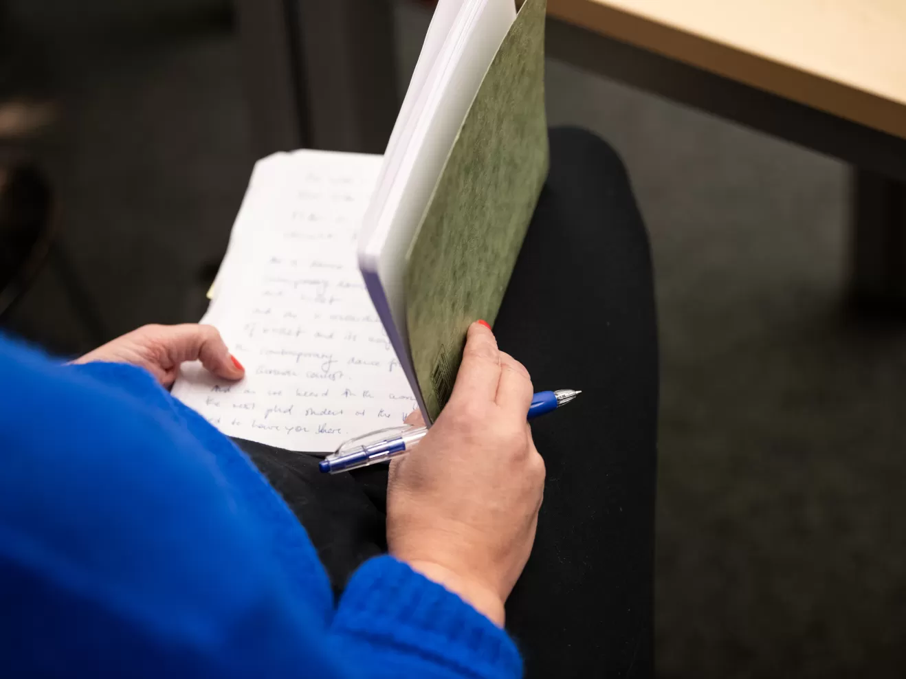 Wir sehen über die rechte Schulter einer sietzenden Person in blauem Pulli auf ein geöffnetes Notizheft und einen Kugelschreiber, die von der Person in Händen gehalten werden.