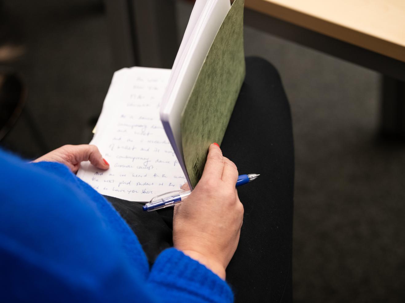 Wir sehen über die rechte Schulter einer sietzenden Person in blauem Pulli auf ein geöffnetes Notizheft und einen Kugelschreiber, die von der Person in Händen gehalten werden.