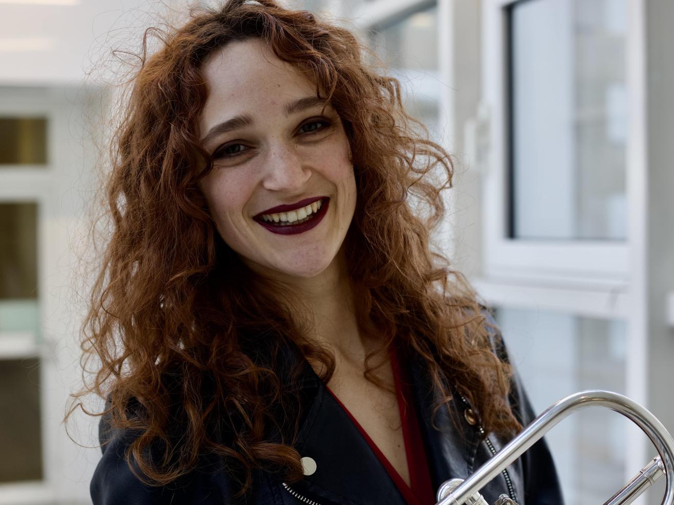 Portrait von Ines Hartmuth: Sie hat rotbraune Locken und lacht in die Kamera. Im Arm hält sie ihre Trompete.