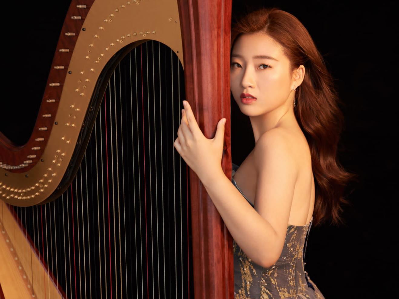 Harfe-Studentin Nan Wang, in einem edlen Kleid, hat die Hand und das Gesicht an der Harfe und blickt ernst in die Kamera.