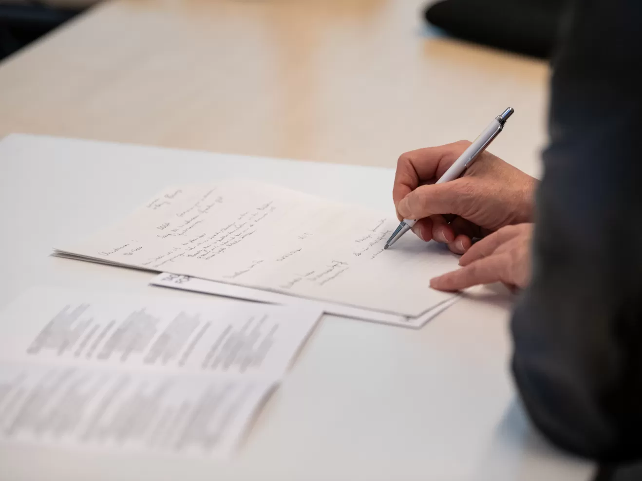 Eine Person macht sich handschriftliche Notizen auf einem Zettel auf einem Tisch, auf dem auch ein Tagungsprogramm aufgeschlagen liegt. Von der Person sind durch den gewählten Bildausschnitt nur die Hände und ein Arm sichtbar.