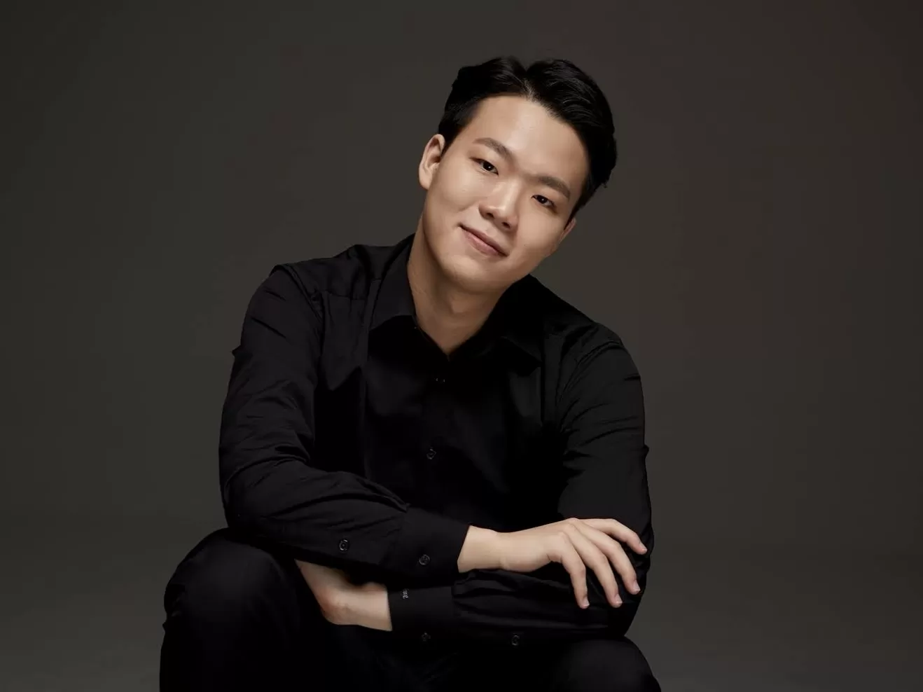 Jaehyun Jeon trägt schicke, schwarze Kleidung und lächelt in die Kamera. Er ist in der Hocke und hat die Arme übereinander gelegt. Der Hintergrund ist grau.