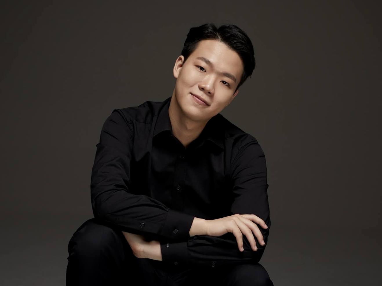 Jaehyun Jeon trägt schicke, schwarze Kleidung und lächelt in die Kamera. Er ist in der Hocke und hat die Arme übereinander gelegt. Der Hintergrund ist grau.