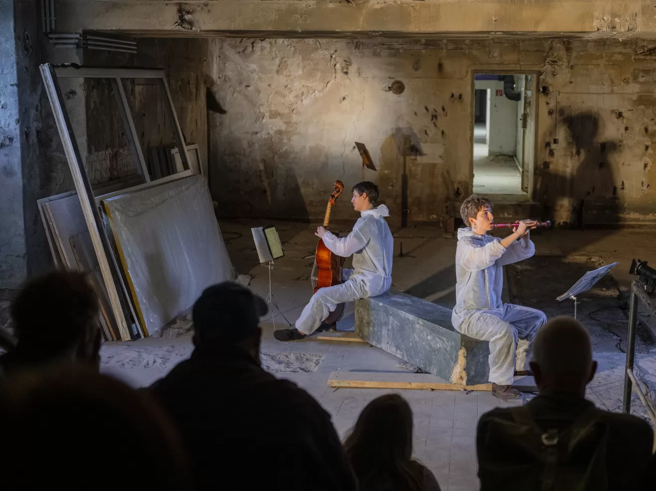 Ein Cellist und eine Blockflötistin sitzen auf einer Art rechteckigem Stein in einem baufällig aussehenden Keller und spielen. Im Anschnitt sind Zuschauer*innen zu sehen.