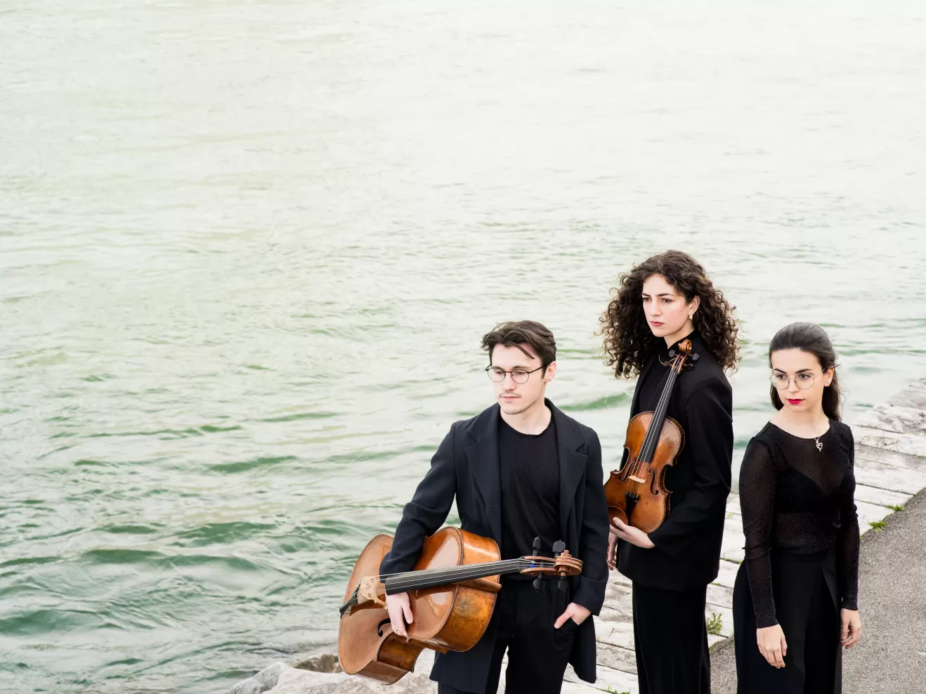 Die drei Musiker*innen des Trio Achai stehen am Hafen, neben ihnen das Meer.