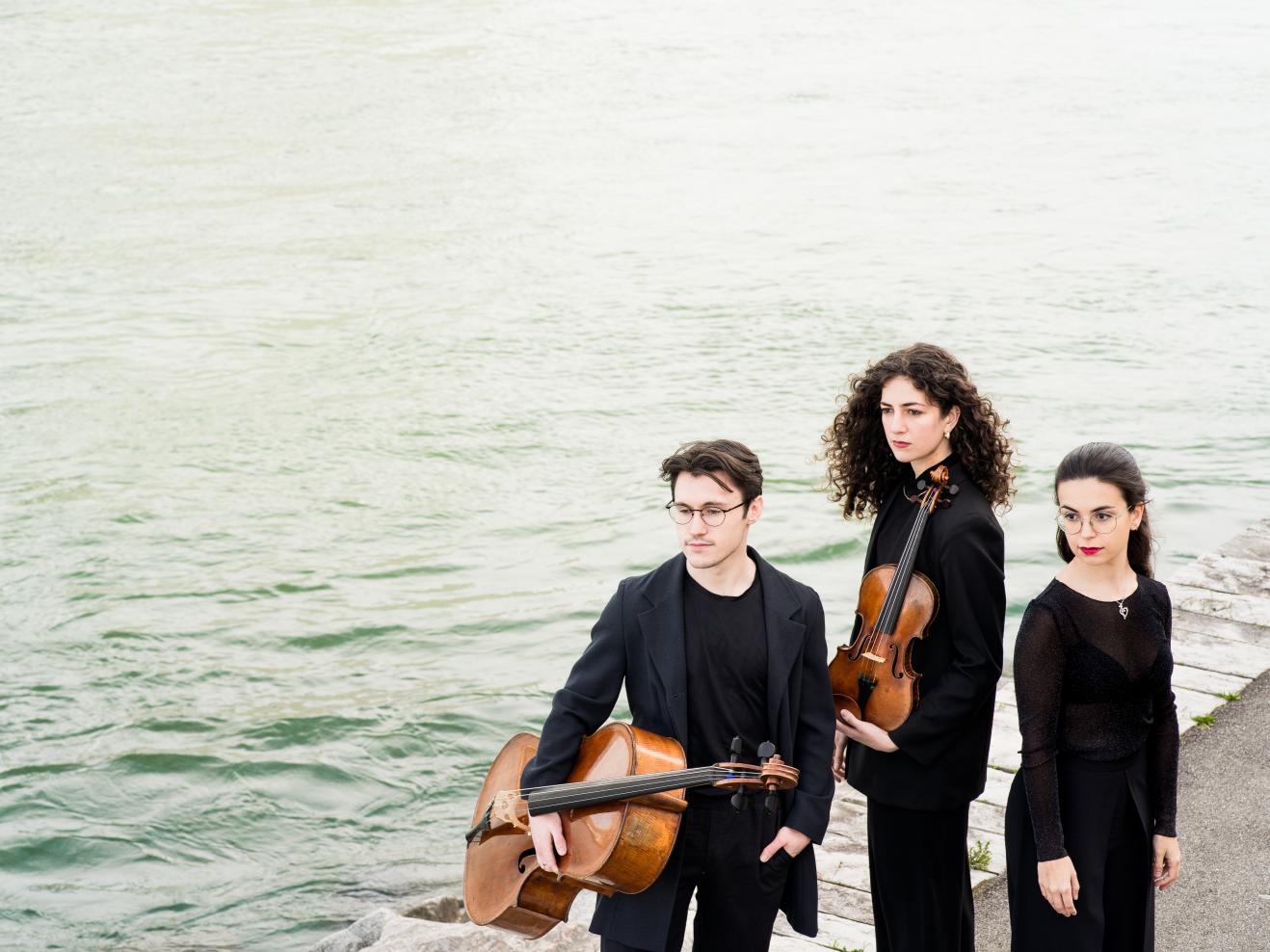 Die drei Musiker*innen des Trio Achai stehen am Hafen, neben ihnen das Meer.